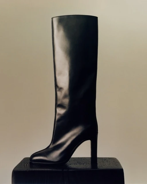 Black knee-high heeled boot displayed on a black wooden block against a neutral backdrop in a studio setting. Fashion photography.