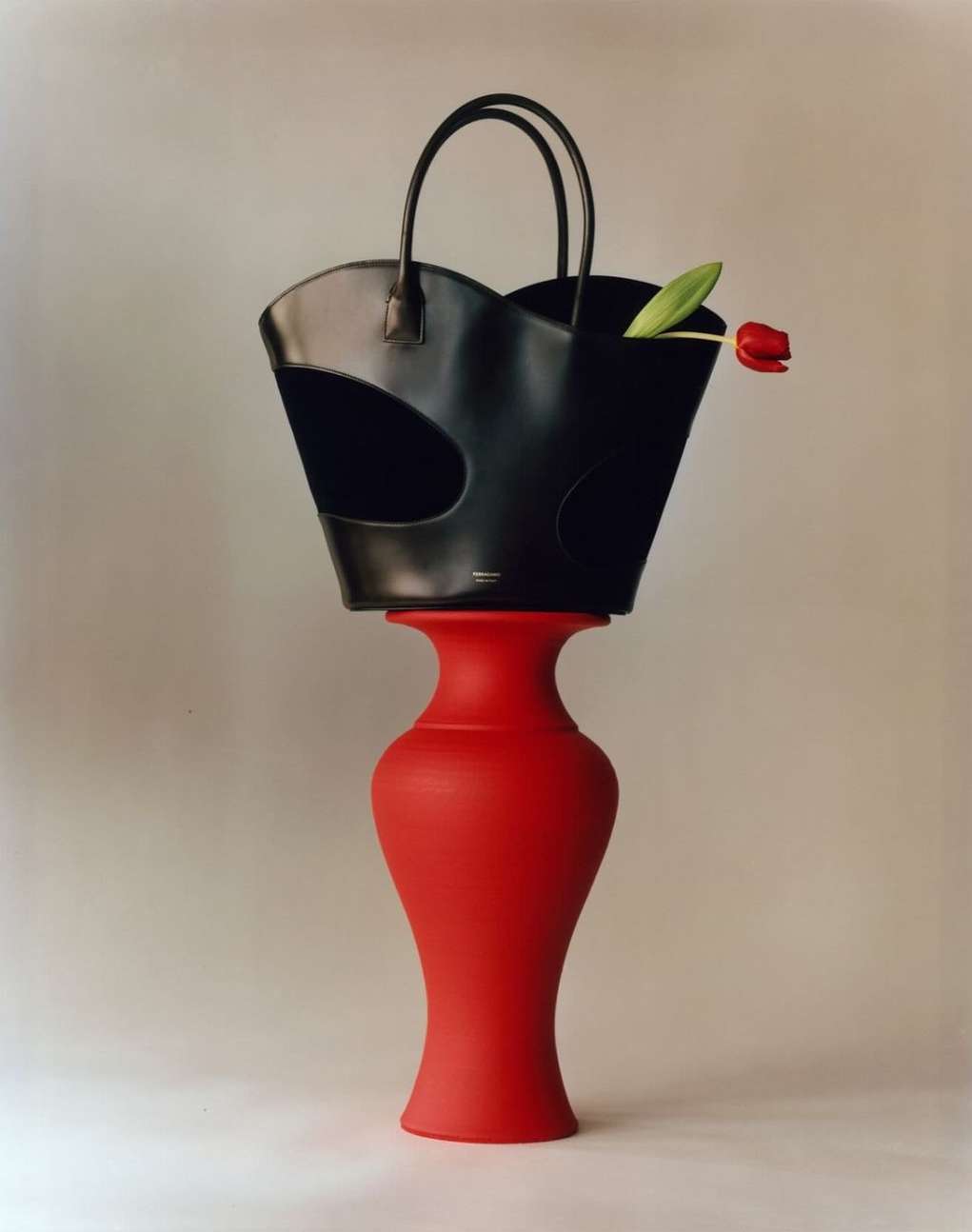 Studio shot of a black handbag on a red vase with a single red tulip, against a light grey background, fashion still life.
