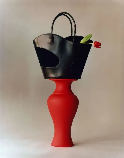 Black handbag with a tulip inside perched atop a red hourglass-shaped stand in a studio setting with even lighting and soft shadows.