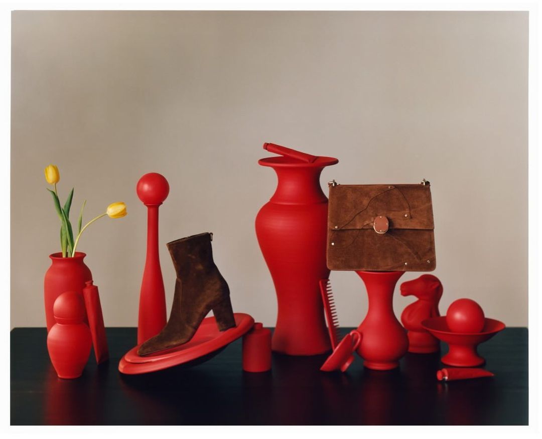 Still life arrangement of a brown heeled boot, handbag, yellow tulips and an array of red vases and decorative objects.