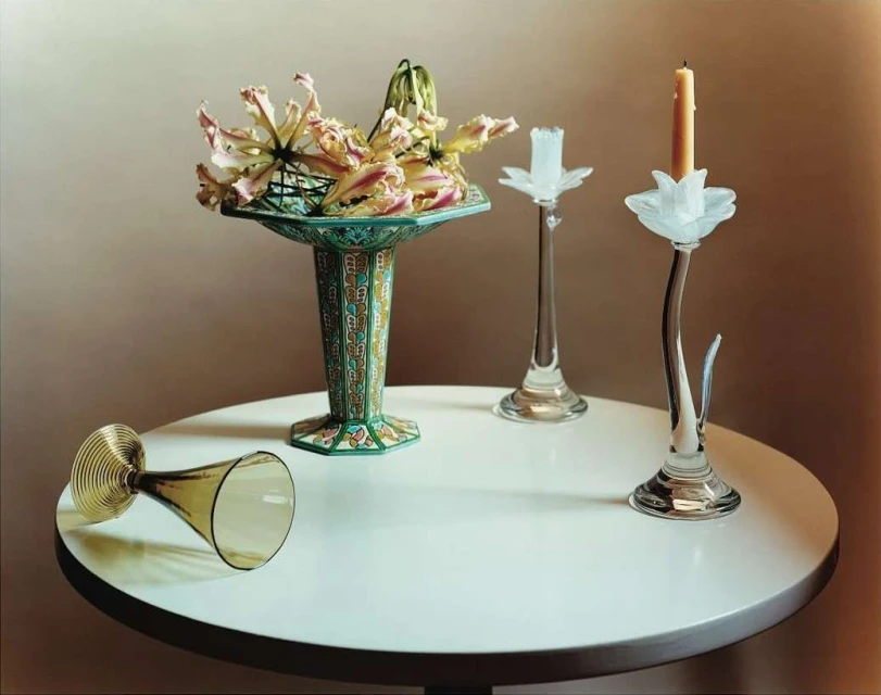 Elegant still life showcases exotic lilies in an ornate vase, alongside a tipped glass and two candle holders on a white table against a neutral backdrop.