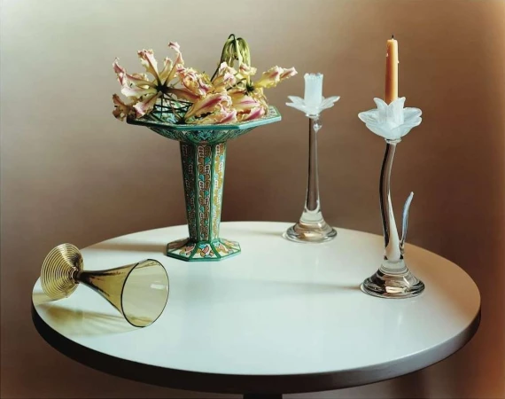 Elegant still life showcases exotic lilies in an ornate vase, alongside a tipped glass and two candle holders on a white table against a neutral backdrop.