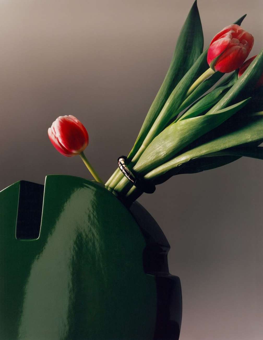 Still life of a green vase holding a bouquet of red and white tulips tied with a dark band, set against a neutral gray background.