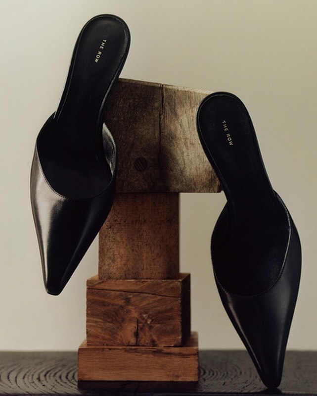 Elevated studio shot of two black leather heeled mules from The Row, set on a stacked wooden base, conveying luxury fashion and style.
