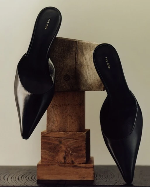Elevated studio shot of two black leather heeled mules from The Row, set on a stacked wooden base, conveying luxury fashion and style.
