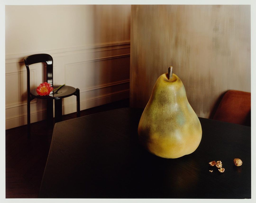 Still life photograph featuring a green and yellow pear on a black table, a red flower on a chair, and neutral-toned walls.