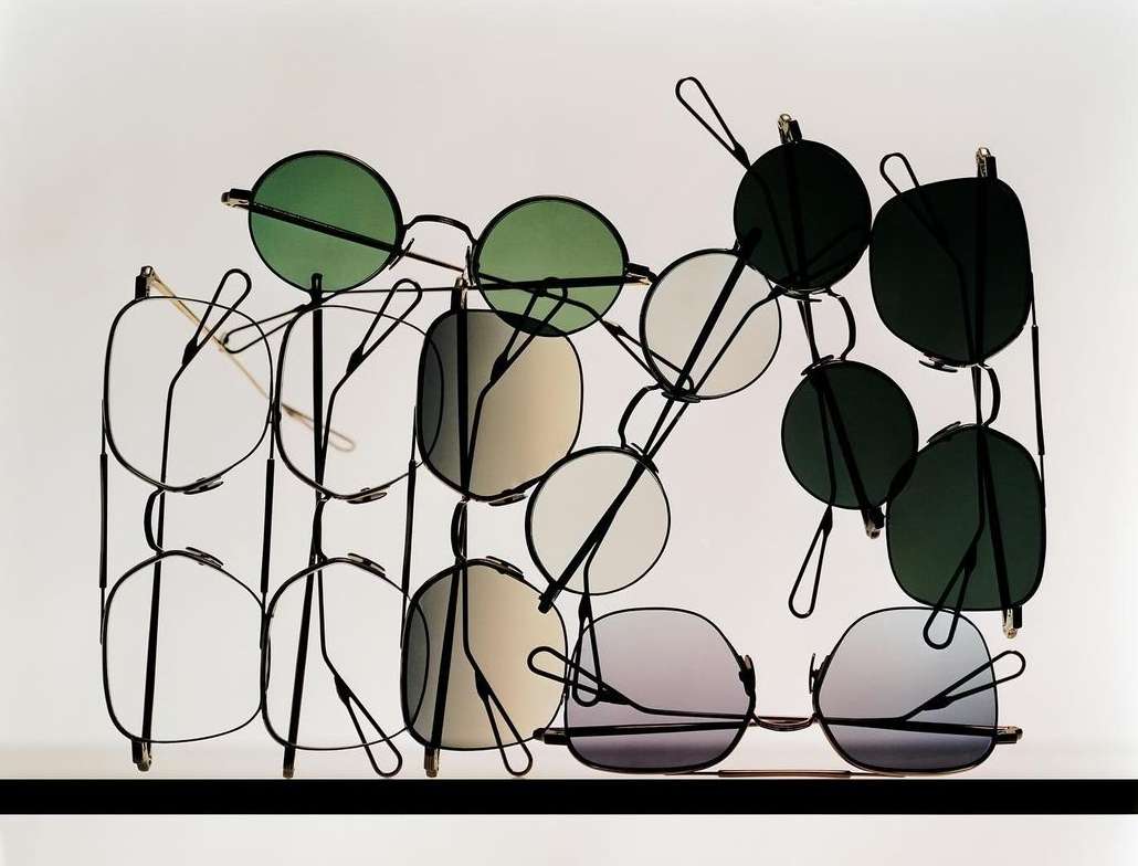 Display of multiple pairs of glasses with various lens colors and frame shapes arranged in a metal rack against a white background.