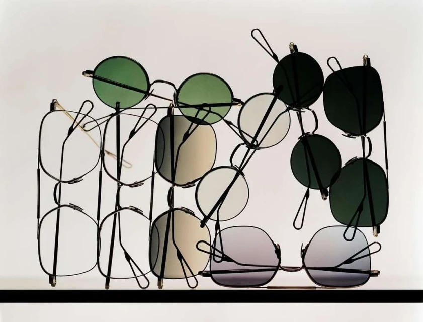 Display of multiple pairs of glasses with various lens colors and frame shapes arranged in a metal rack against a white background.