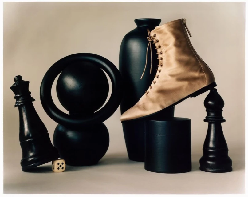 Beige lace-up boot balances on a black bishop chess piece, surrounded by a vase, black ring sculpture, cylinder, die, and a chess queen.