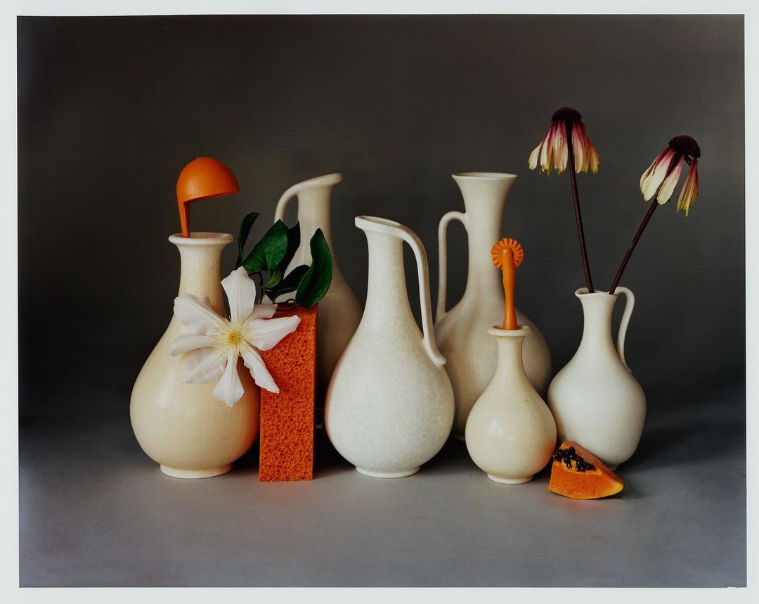 Still life with white vases holding flowers and decorative objects, set against a dark gray backdrop in a studio setting.