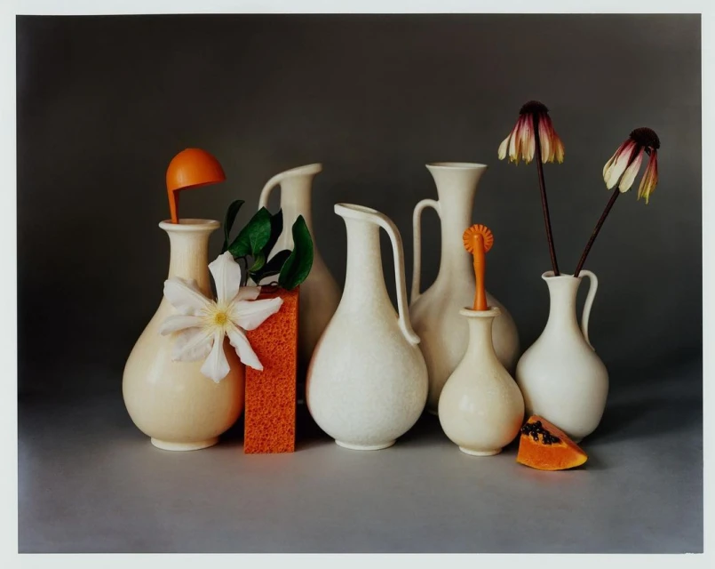 Still life with white vases holding flowers and decorative objects, set against a dark gray backdrop in a studio setting.