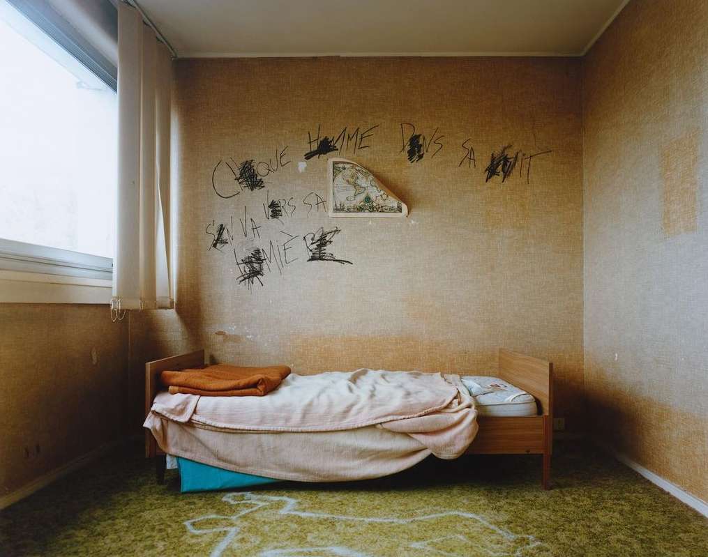 Bedroom interior featuring a bed with a blanket, patterned wallpaper, and graffiti-like writing on the wall, creating an eerie atmosphere.