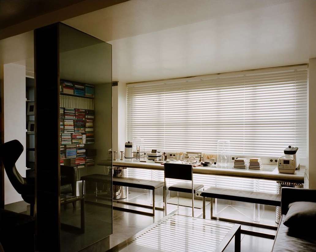 Interior shot of a room with a wall of windows, a table with objects, bookshelves, and furniture, creating a modern and minimalist aesthetic.
