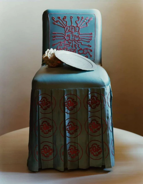 Still life featuring a blue chair with a decorated back and skirt, topped with a plate and cream, set against a plain backdrop.