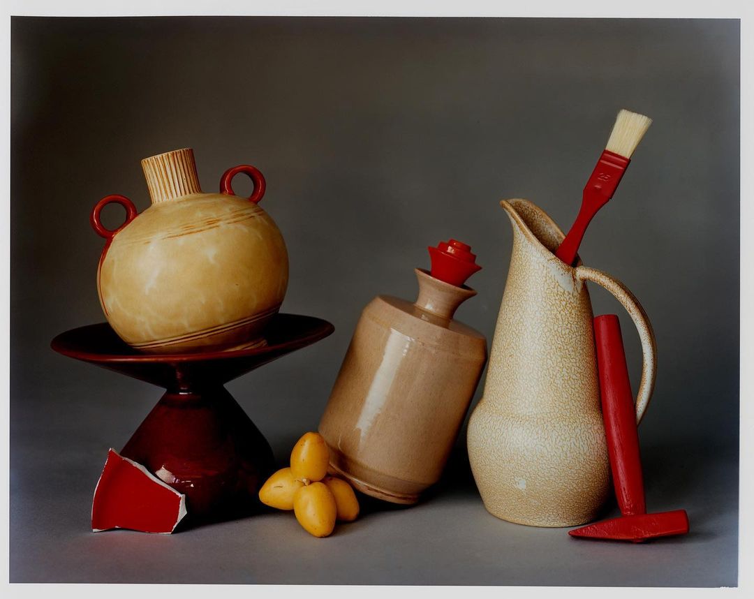 Still life photograph featuring ceramic vases, a red paintbrush, and a few dates against a grey backdrop in an indoor setting.