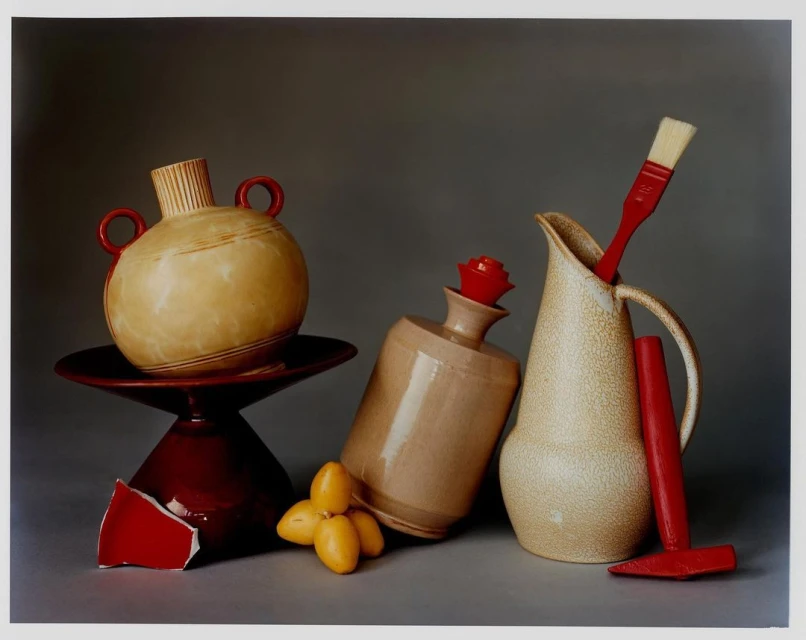 Still life photograph featuring ceramic vases, a red paintbrush, and a few dates against a grey backdrop in an indoor setting.