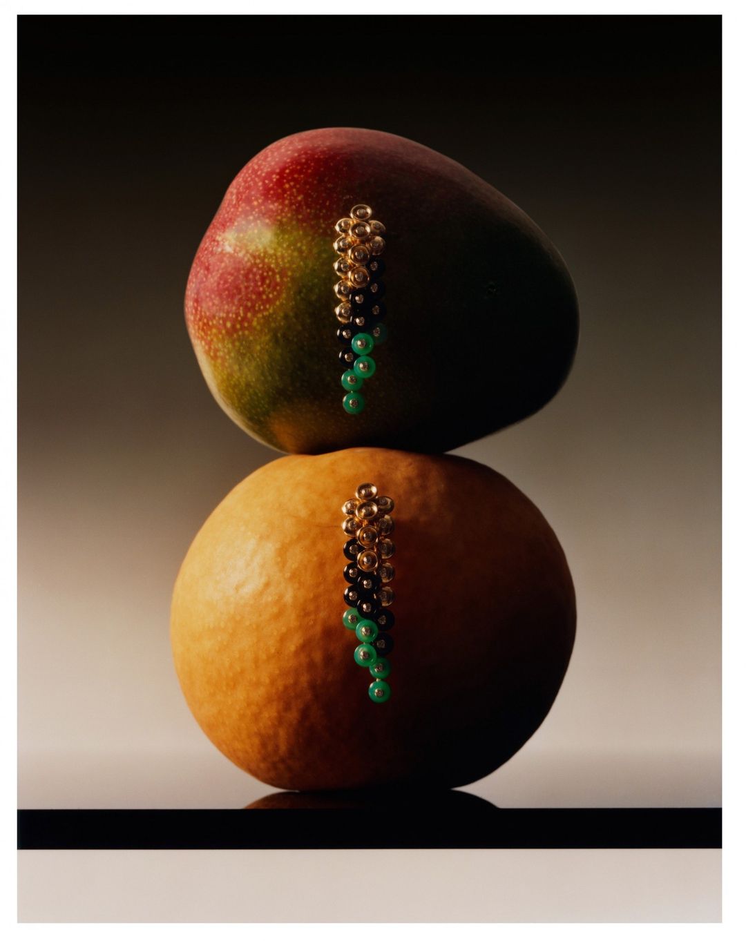 Still life studio shot of a mango and an orange stacked, each adorned with a dangling gold and green beaded earring. 