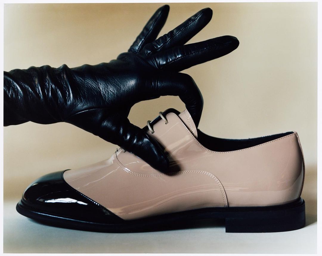 Patent leather dress shoe held by a black leather glove against a light neutral background, with shiny highlights, shadows, and textures.