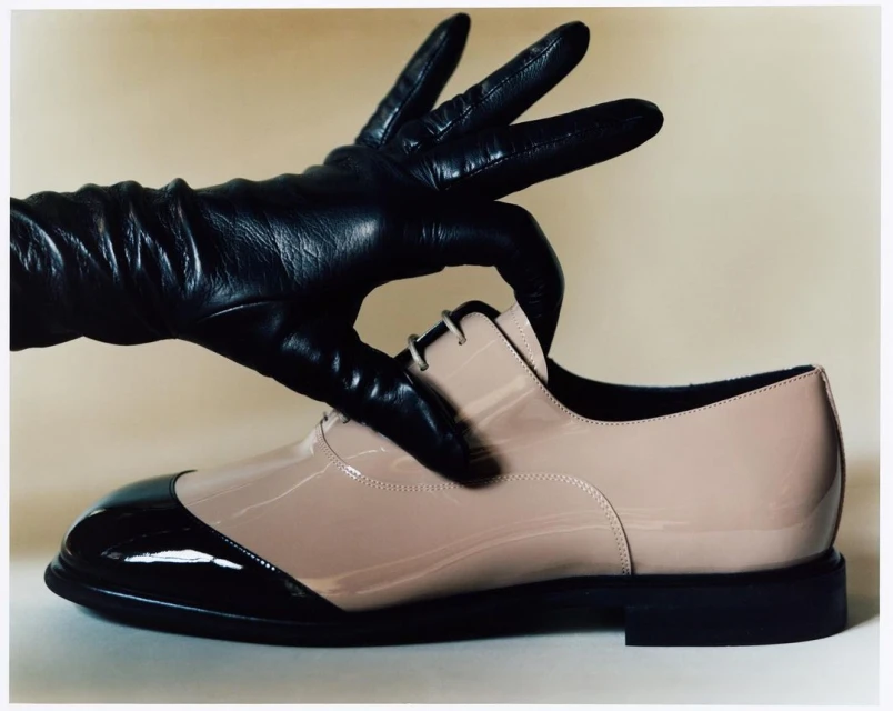Patent leather dress shoe held by a black leather glove against a light neutral background, with shiny highlights, shadows, and textures.