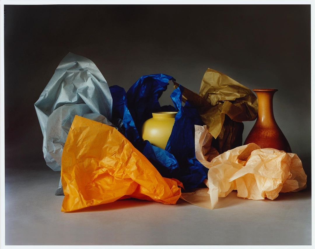 Still life photograph featuring a yellow vase surrounded by crumpled sheets of colorful paper, set against a grey backdrop with soft, diffused lighting.