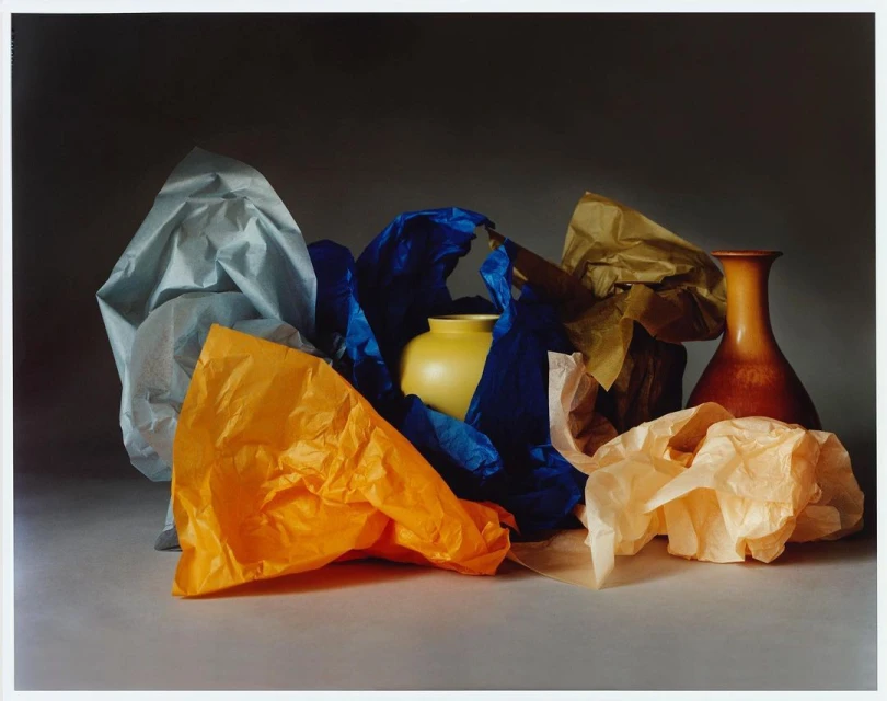 Still life photograph featuring a yellow vase surrounded by crumpled sheets of colorful paper, set against a grey backdrop with soft, diffused lighting.