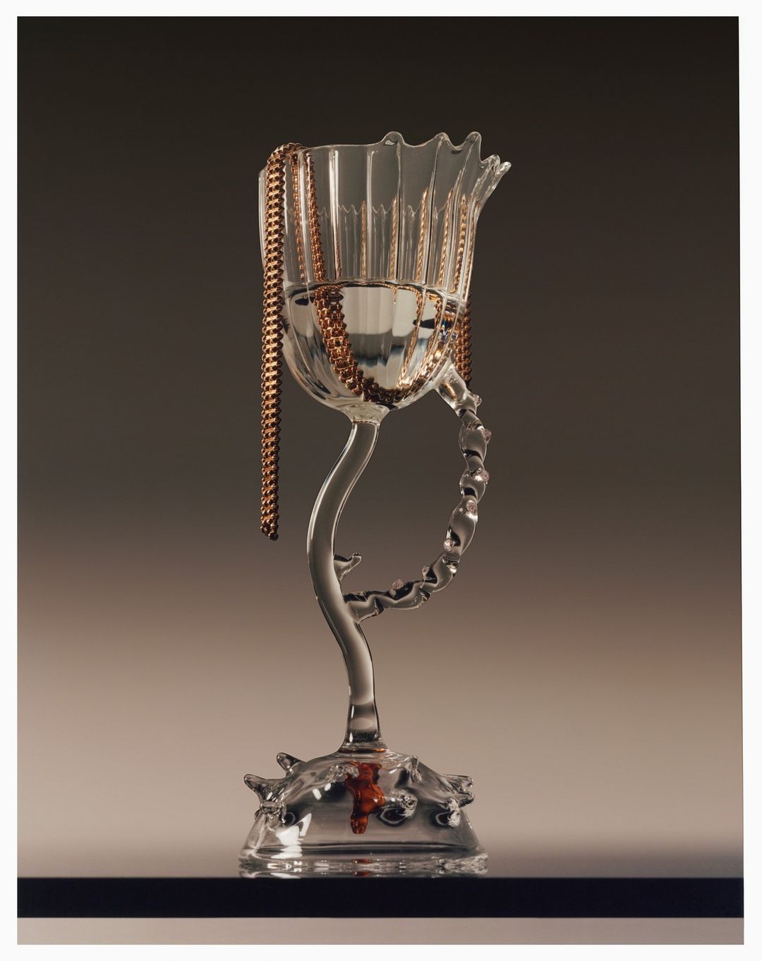 A transparent glass chalice with a stem features a copper chain draped over it against a gradient grey background, high key studio lighting.