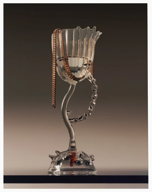 A transparent glass chalice with a stem features a copper chain draped over it against a gradient grey background, high key studio lighting.