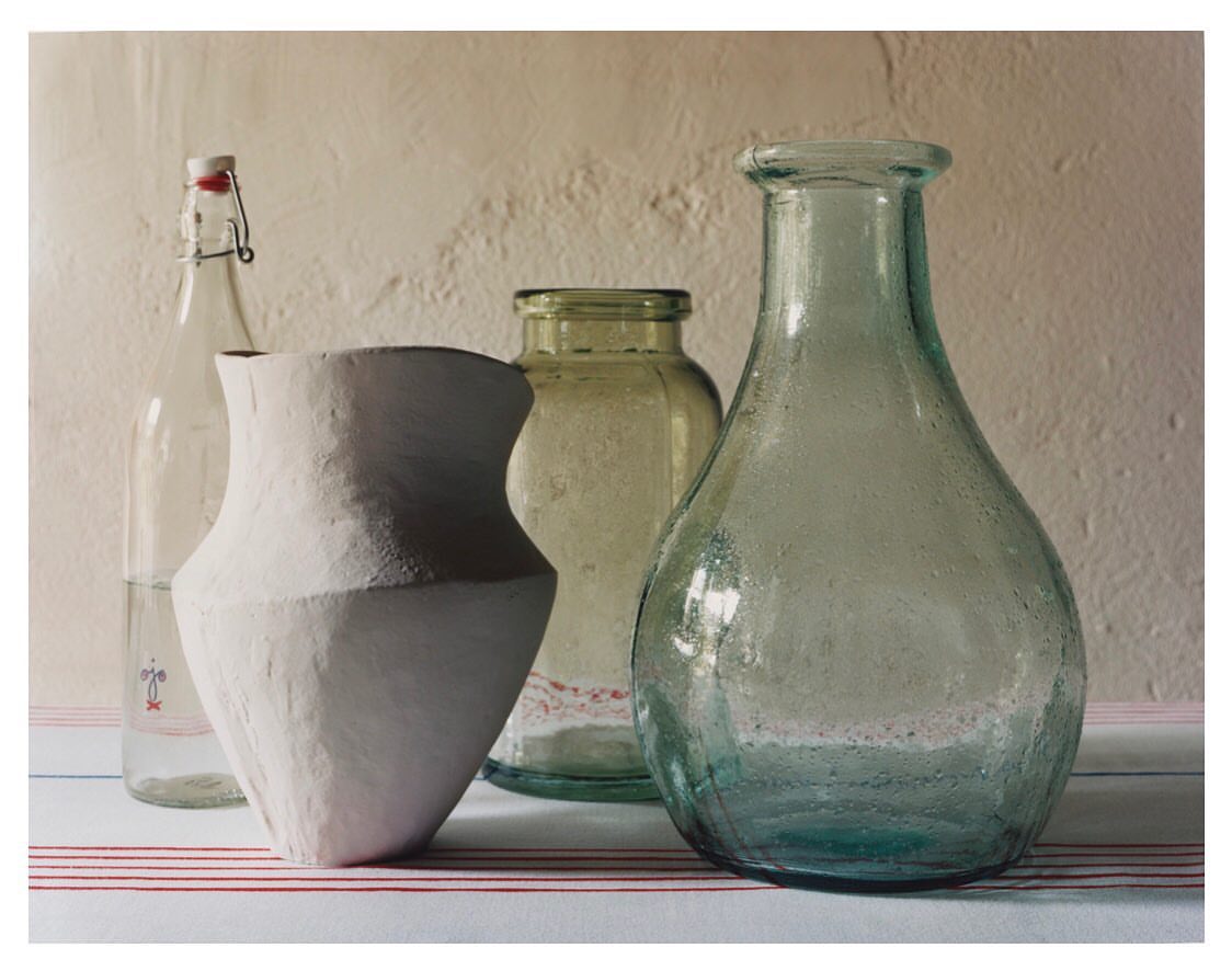 Still life photograph of an arrangement of clear glass bottles and a light-colored ceramic vase on a striped tablecloth.