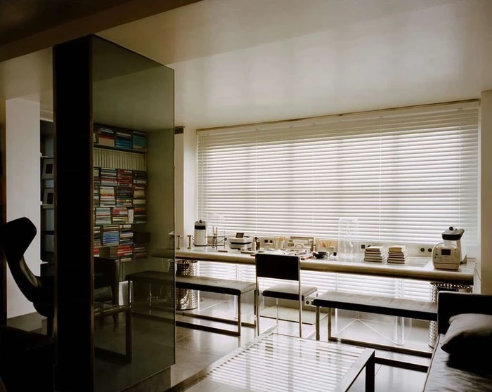 Interior shot of a modern minimalist living room with a long desk, white window blinds, and a mirrored glass divider column