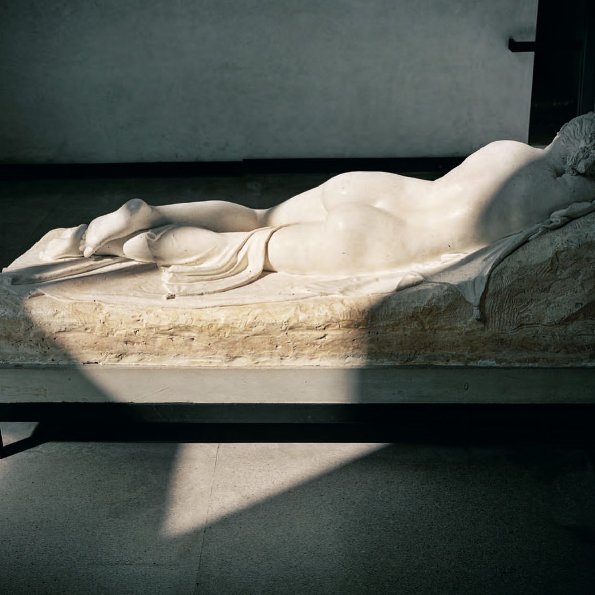 Marble sculpture of a reclining nude figure indoors with light and shadow play on a platform in a museum setting, classical art piece.