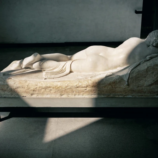 Marble sculpture of a reclining nude figure indoors with light and shadow play on a platform in a museum setting, classical art piece.