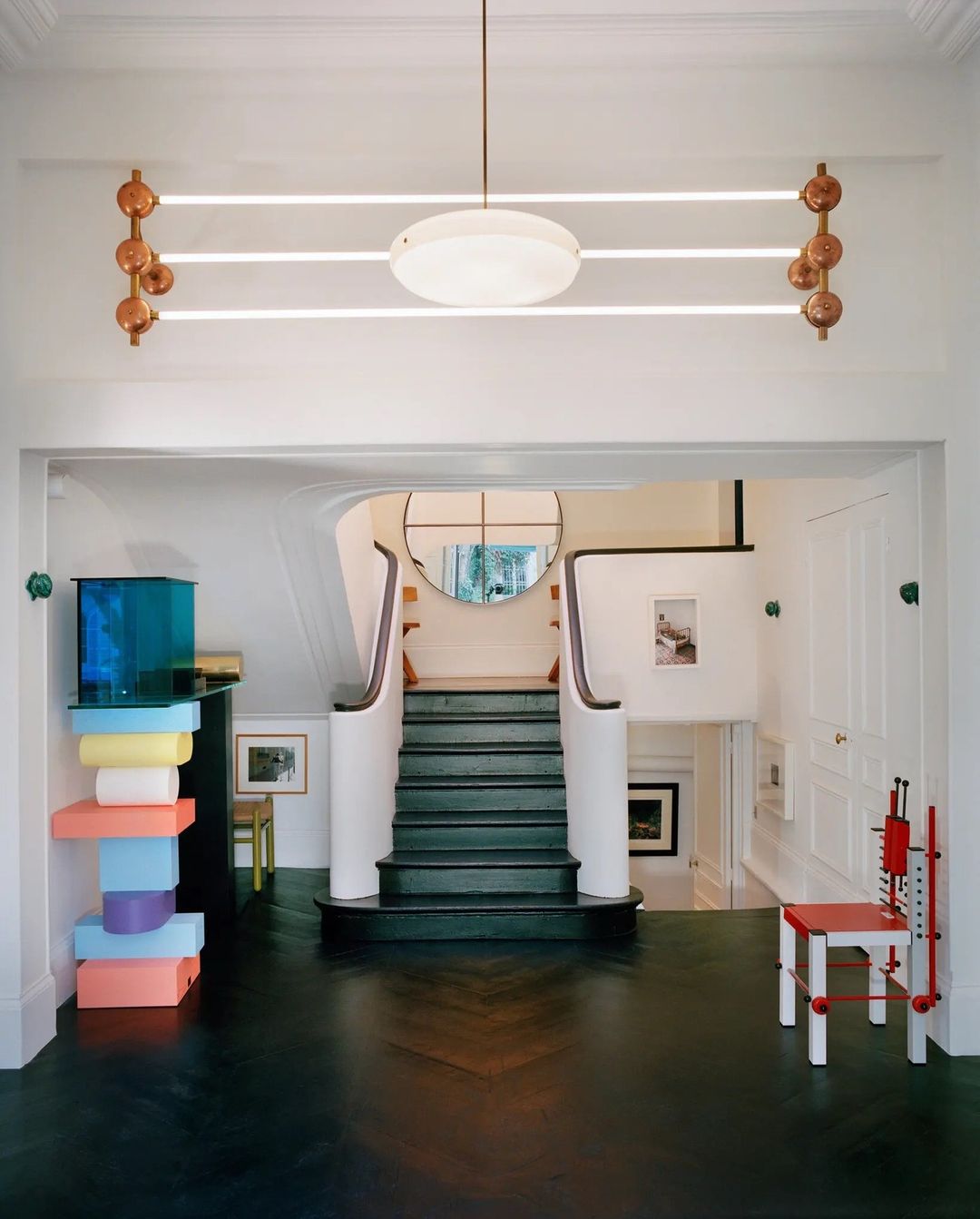 Interior view of a home hallway with a staircase, custom lighting, and colorful art and furniture under bright, natural lighting.