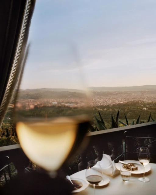 A glass of white wine in the foreground, framing a distant cityscape and a table setting with plates and glassware in the background.