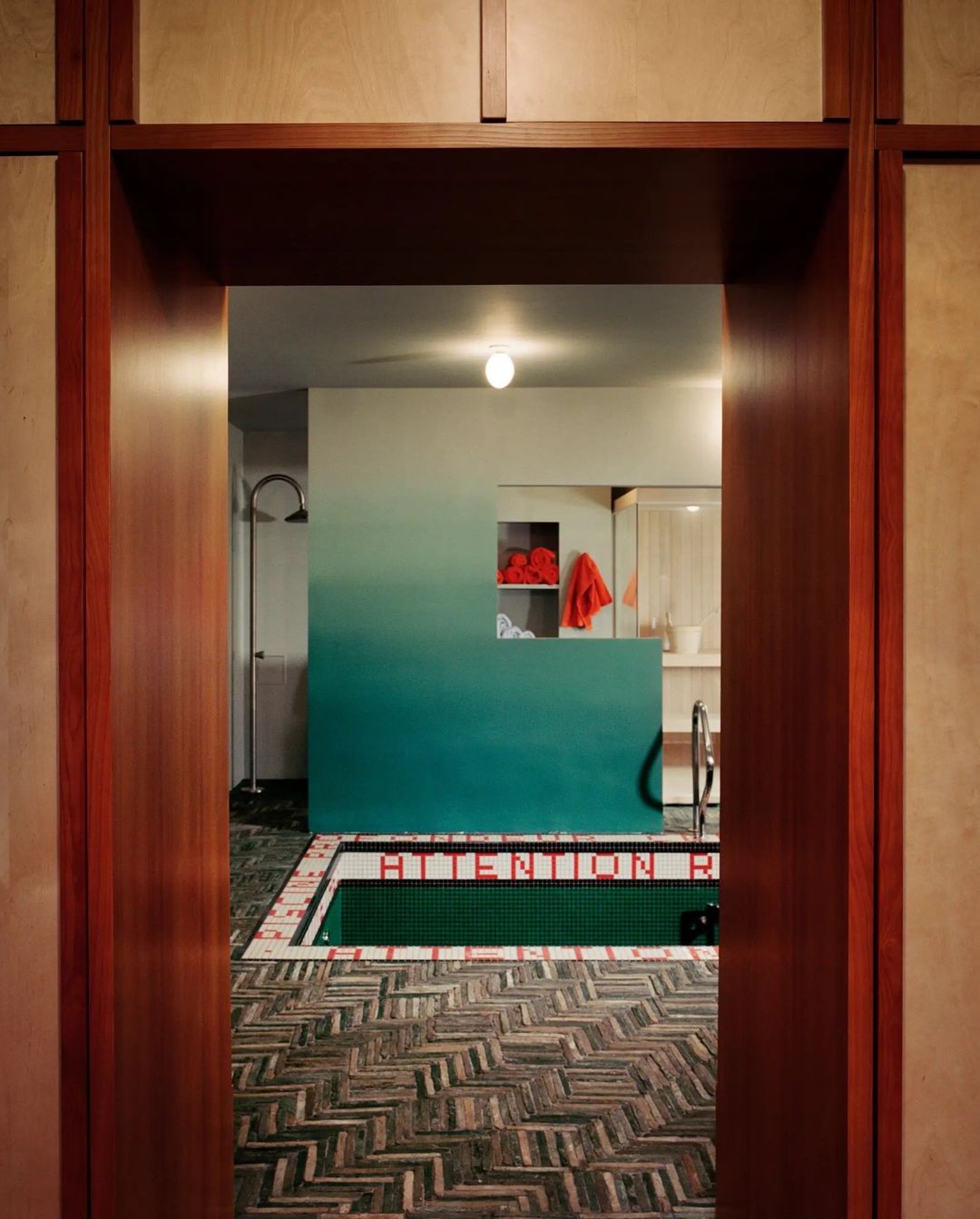 Interior view of indoor pool with green tile and herringbone floor through paneled wood doorway; red towels in recessed wall shelf.