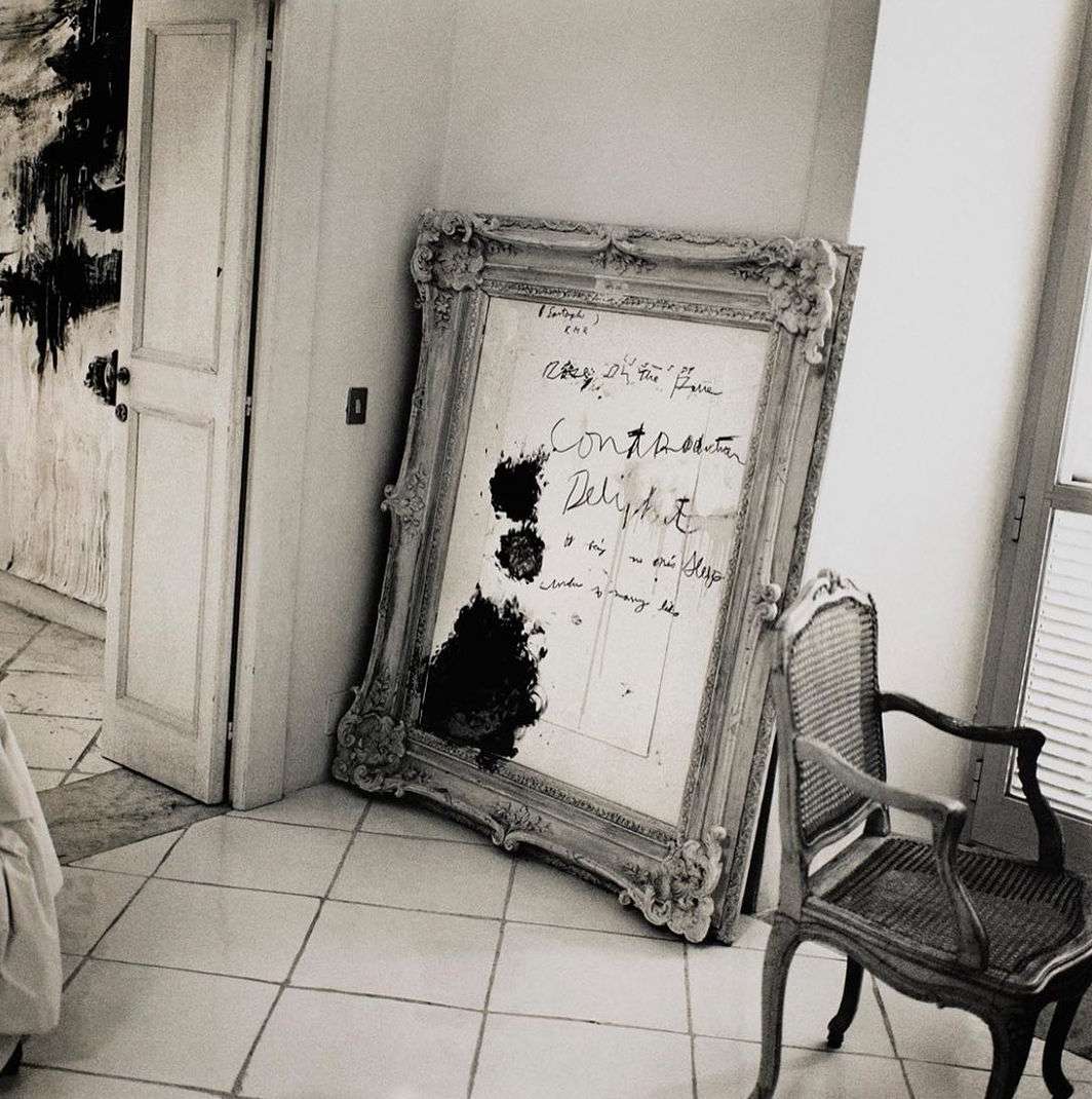 Vintage-style photo of a leaning ornate mirror with handwritten text and smudges, alongside a cane chair, on a tiled floor near a door.