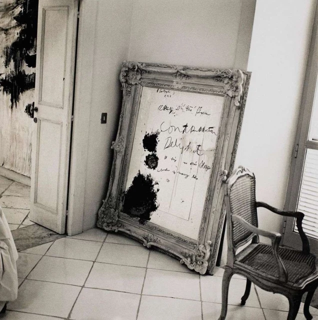 Vintage-style photo of a leaning ornate mirror with handwritten text and smudges, alongside a cane chair, on a tiled floor near a door.