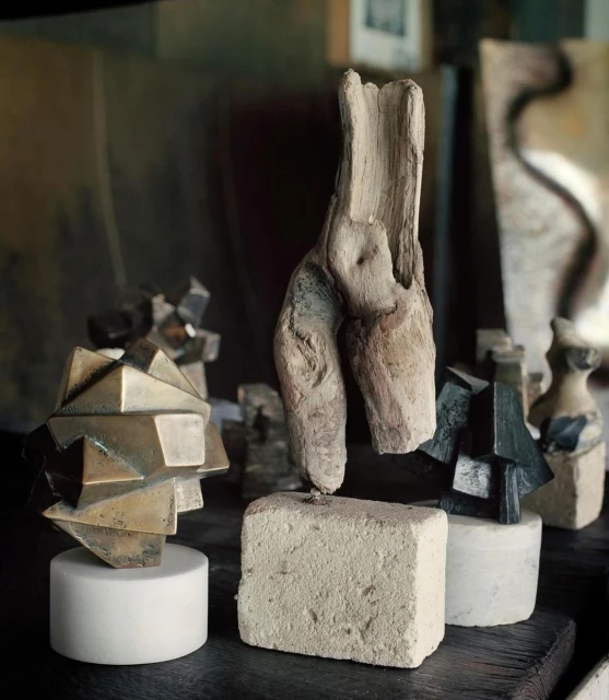 Art studio scene featuring a collection of geometric and organic sculptures displayed on blocks and tables, lit with ambient light.