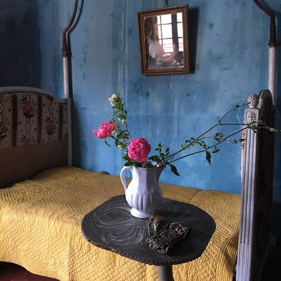 Interior still life of a vintage bed with a yellow cover, a small round table holding flowers in a white pitcher, and a framed picture on a textured blue wall.