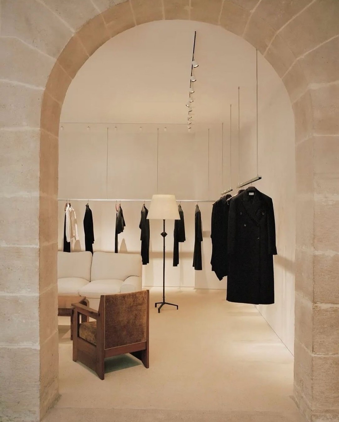 Clothing store interior with minimalist design, featuring racks of apparel, a sofa, lamp, and chair under neutral, indirect lighting.