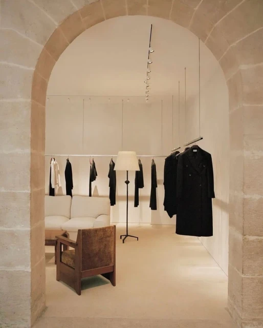 Clothing store interior with minimalist design, featuring racks of apparel, a sofa, lamp, and chair under neutral, indirect lighting.
