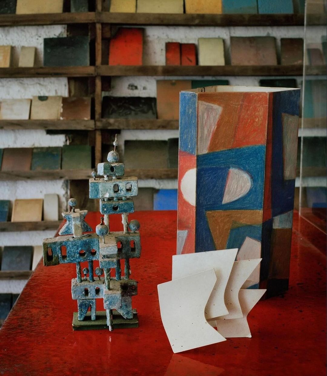 Abstract arrangement featuring a geometric sculpture, painted geometric box, and curved paper on a glossy red surface with tile shelves.