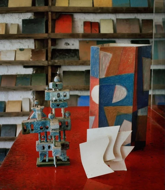 Abstract arrangement featuring a geometric sculpture, painted geometric box, and curved paper on a glossy red surface with tile shelves.