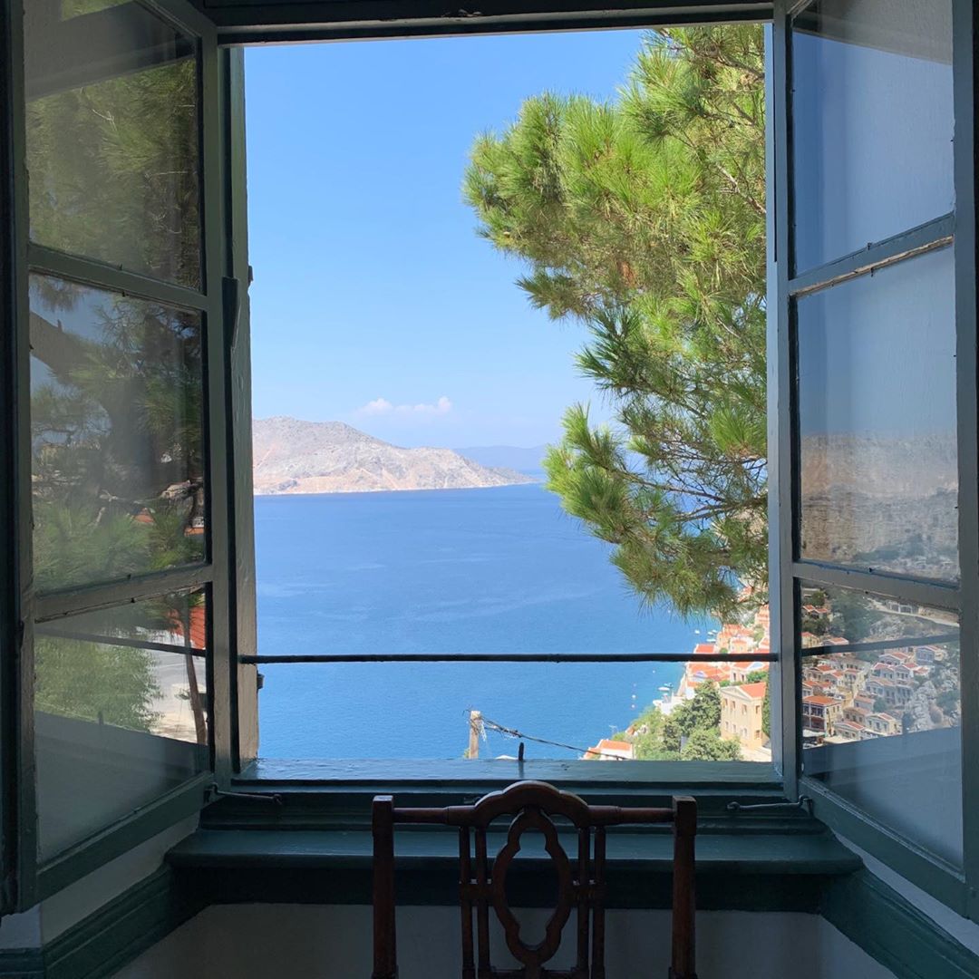 Open window view of the ocean and distant hills, bright sunny day, framed by a wooden chair and architecture details.