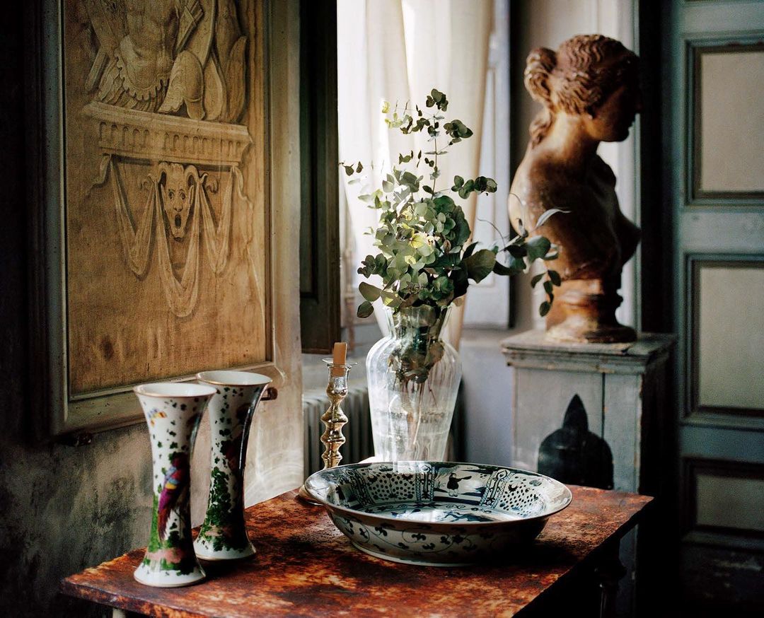 Ornate interior scene featuring a brown table with floral vases, an antique bowl, and a bust sculpture against a window backdrop.