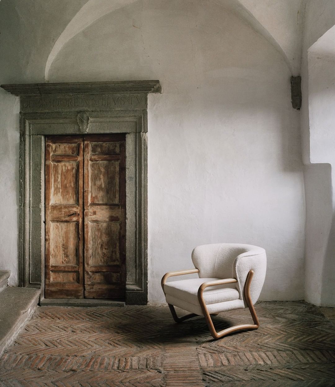 Interior view of a cream-colored armchair with a wooden frame on a brick floor, set against a white wall with an arched doorway and a closed wooden door.