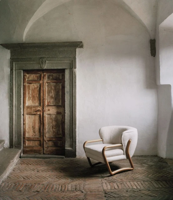 Interior view of a cream-colored armchair with a wooden frame on a brick floor, set against a white wall with an arched doorway and a closed wooden door.