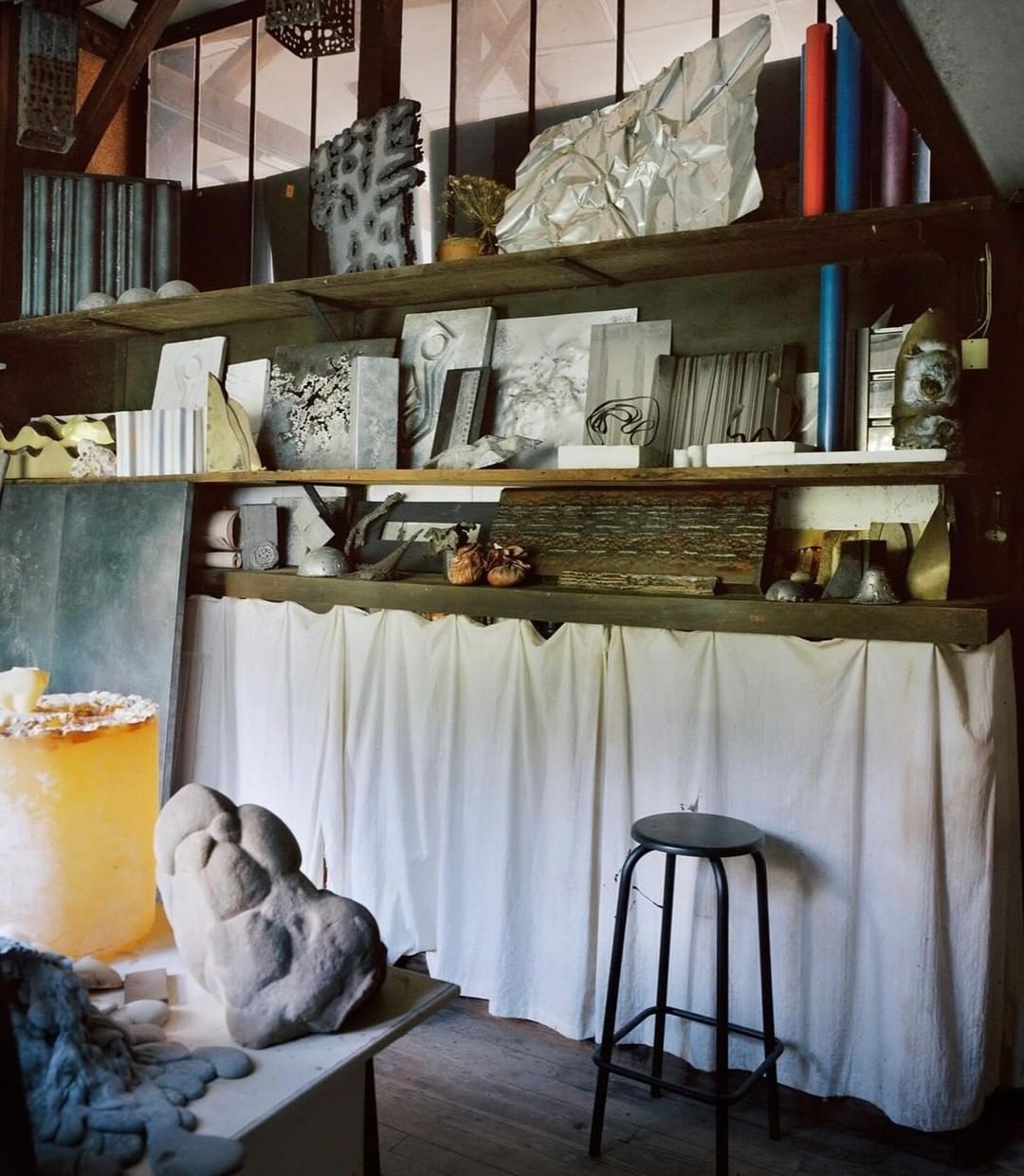Art studio interior filled with sculptures and artwork on shelves, bathed in natural light with wooden beams and a stool.