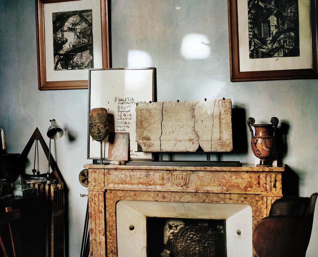 Interior view with a marble fireplace topped with a stone fragment with inscription, a metal head sculpture, and various decorative objects.