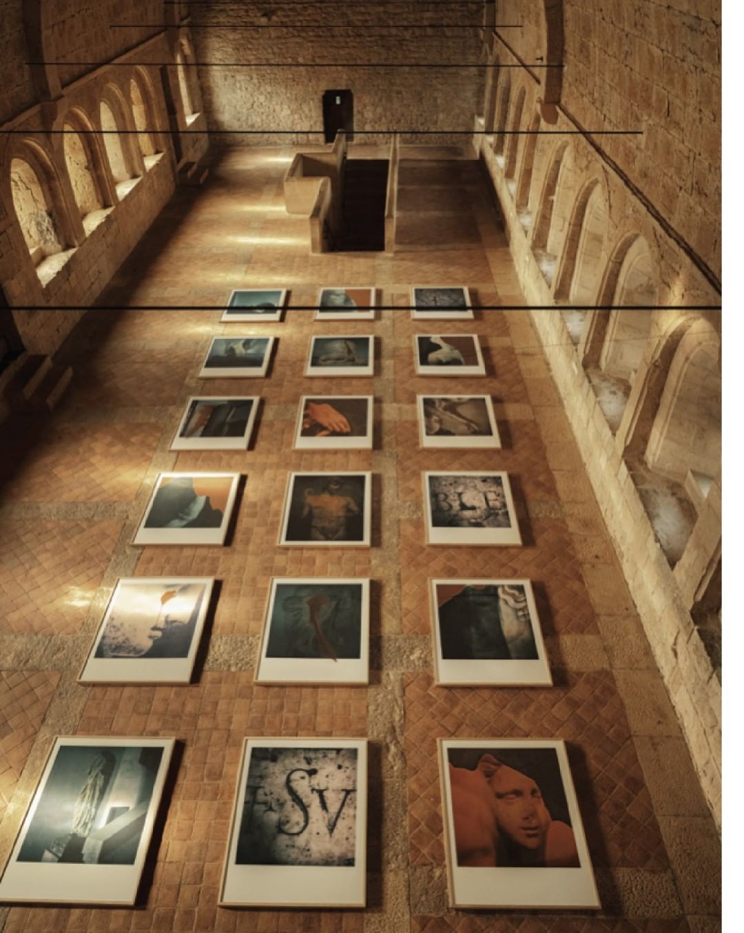 Indoor art gallery with a series of framed pictures hanging on a brick wall under arched windows, creating a visual display.