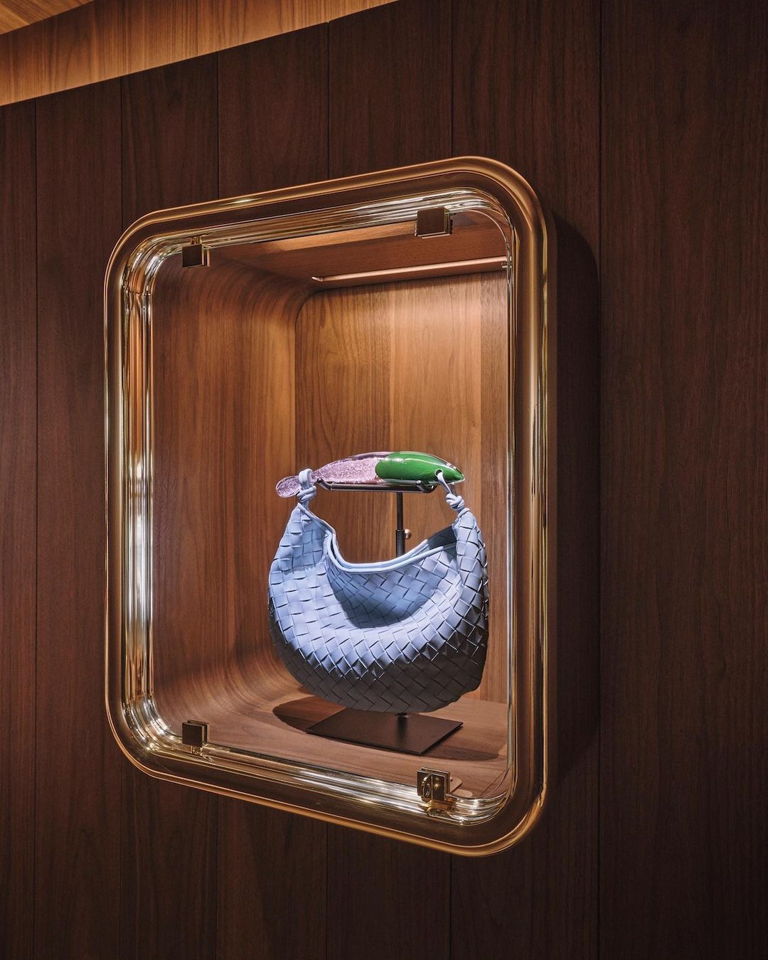 Fashionable blue Bottega Veneta purse on a display stand inside a wall-mounted, gold-trimmed, glass display case with wood panelling.